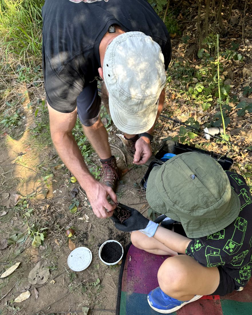 An adult and child crouched together outdoors, threading a worm onto a fishing line in the Queensland bush